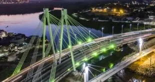 Ponte estaiada iluminada de verde à noite sobre o rio, com a cidade ao fundo