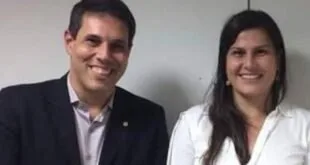 Homem de terno e mulher de camisa branca sorriem lado a lado em ambiente interno