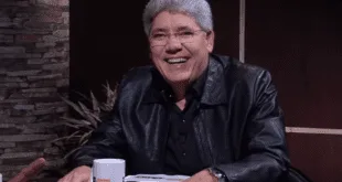 Homem de cabelos grisalhos e jaqueta de couro preta sorrindo sentado à mesa com um livro