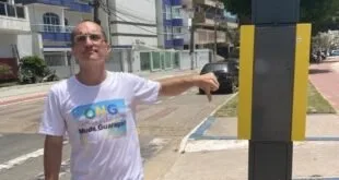 Homem de camiseta branca apontando para um parquímetro na calçada, com prédios ao fundo