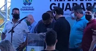 Pessoas reunidas em torno de um púlpito diante de painel com o brasão e o nome da Prefeitura de Guarapari