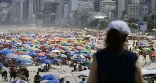 Praia lotada de guarda-sóis coloridos com prédios ao fundo e mulher de chapéu em primeiro plano