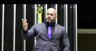 Homem de terno e barba gesticulando na tribuna de um plenário com a bandeira do Brasil ao fundo