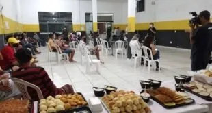 Pessoas sentadas em cadeiras de plástico em um salão, com mesa de salgados em primeiro plano