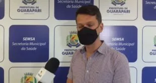 Homem de máscara dando entrevista diante de painel da Secretaria Municipal da Saúde de Guarapari