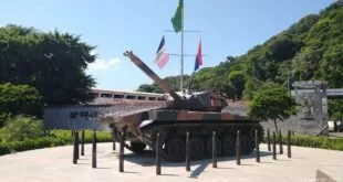 Tanque de guerra exposto em praça com mastros de bandeiras e morro arborizado ao fundo