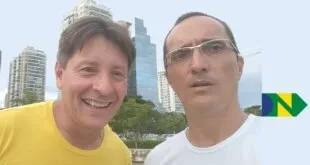 Dois homens sorrindo em selfie ao ar livre com prédios e árvores ao fundo