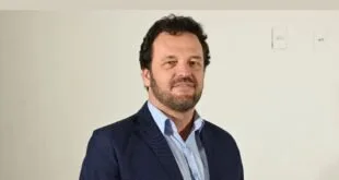 Retrato de homem de barba e blazer escuro posando diante de parede branca