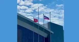 Mastros no alto de um prédio com duas bandeiras hasteadas e um mastro vazio sob céu azul