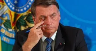 Homem de terno leva a mão ao rosto durante coletiva, com bandeiras desfocadas ao fundo