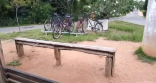 Bicicletas estacionadas em suporte de madeira ao ar livre, com banco rústico e chão de terra