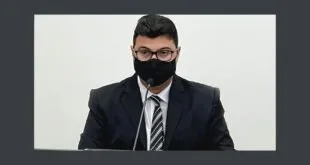 Homem de terno, óculos e máscara preta sentado à mesa em ambiente escuro