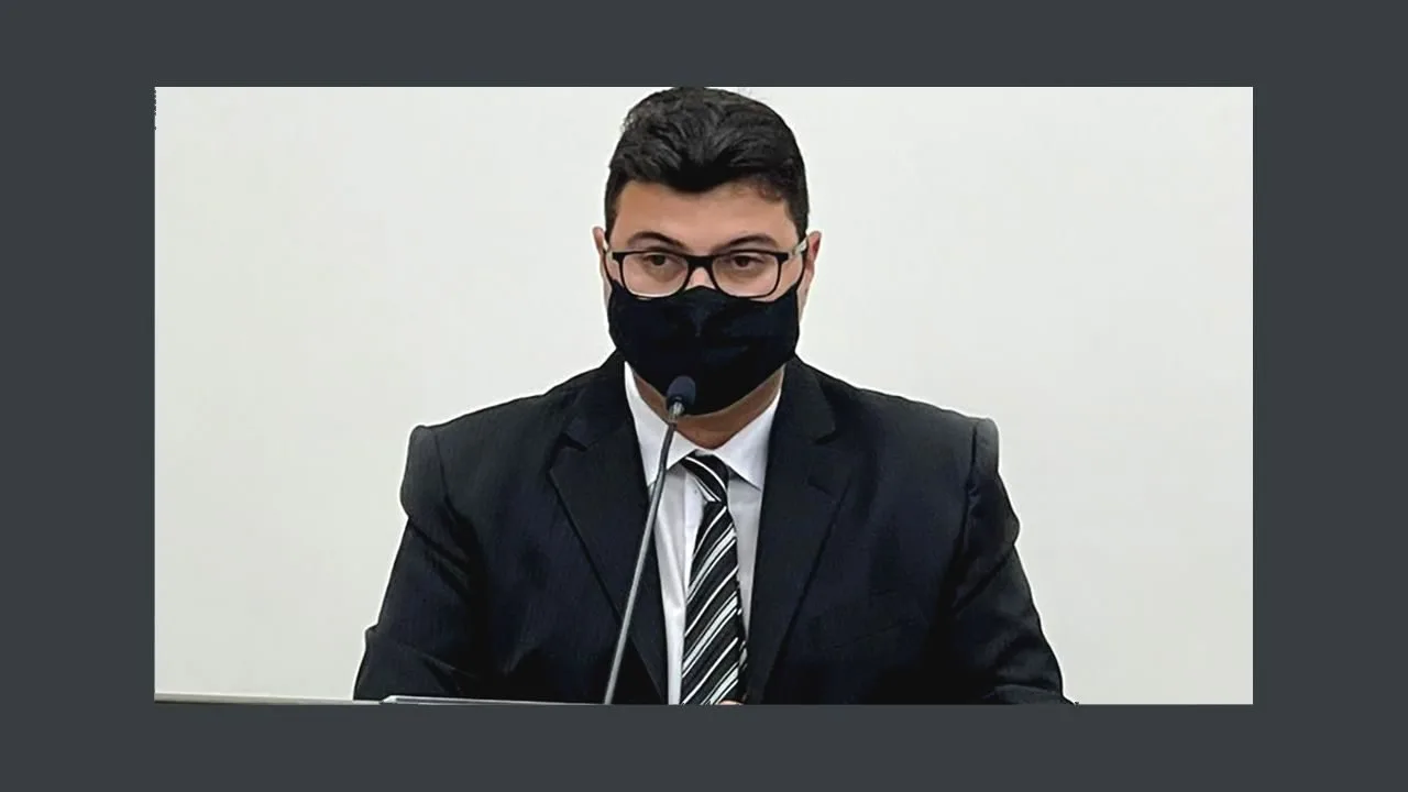 Homem de terno, óculos e máscara preta sentado à mesa em ambiente escuro