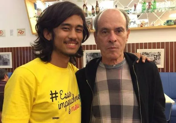 Dois homens posam lado a lado, um deles com camiseta amarela estampada