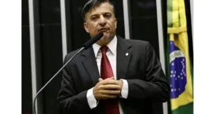 Homem de terno e gravata vermelha discursa em tribuna com bandeira do Brasil ao fundo