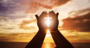 Mãos erguidas em oração formando o desenho de uma cruz contra o sol no fim da tarde