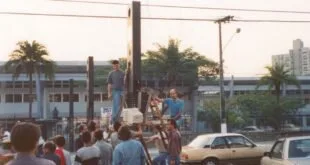 Foto antiga de trabalhadores instalando poste em rua com carros e pessoas ao redor