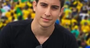 Jovem de camiseta preta posa em manifestação com bandeiras do Brasil ao fundo