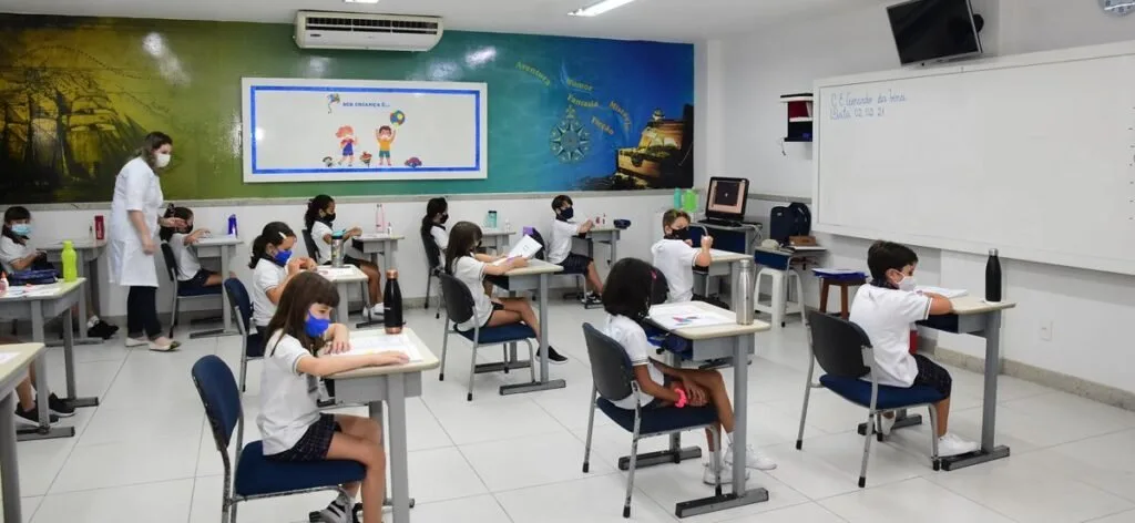 Alunos de uniforme e máscara sentados em carteiras de sala de aula com lousa digital