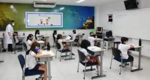 Alunos de uniforme e máscara sentados em carteiras de sala de aula com lousa digital