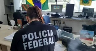 Agente com colete da Polícia Federal diante de computador, com bandeira do Brasil ao fundo
