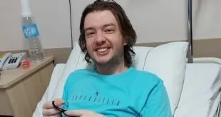 Jovem de camiseta azul sorri sentado em cama de hospital