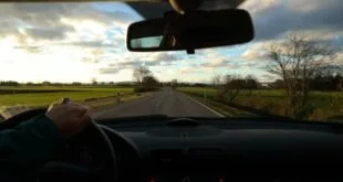 Vista do interior de um carro em estrada, com a mão do motorista no volante