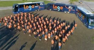 Funcionários de uniforme laranja em formação diante de caminhões com a marca Loga