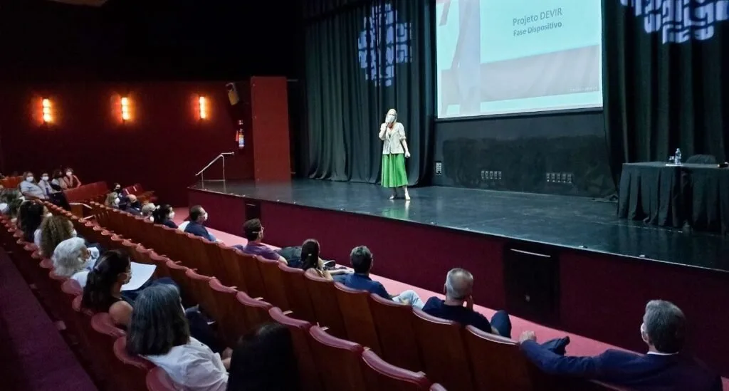 Palestrante de vestido verde no palco de um auditório diante da plateia sentada