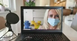 Notebook sobre mesa exibindo videochamada com mulher de máscara, ao lado de microfone