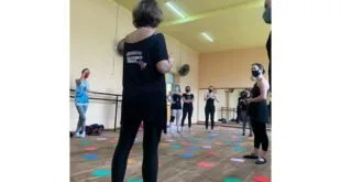 Mulheres de máscara em sala de dança com formas coloridas espalhadas pelo piso de madeira