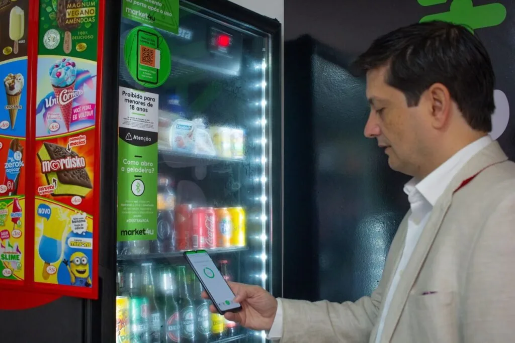 Homem de blazer claro usa o celular diante de geladeira iluminada de mercado autônomo