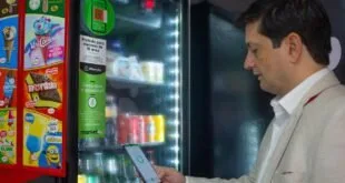 Homem de blazer claro usa o celular diante de geladeira iluminada de mercado autônomo