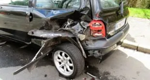 Carro preto com a traseira destruída após colisão, parado à beira da via