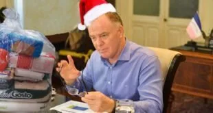 Homem de camisa azul e gorro de Papai Noel gesticula sentado à mesa de reunião