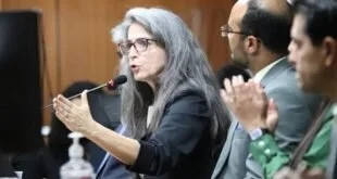 Mulher de óculos gesticula ao falar em mesa de evento enquanto homem ao lado aplaude