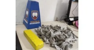 Tablete amarelo e dezenas de embalagens de droga sobre mesa ao lado de placa da Polícia Militar