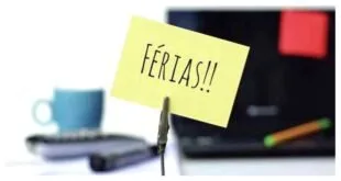 Bilhete amarelo escrito 'FÉRIAS!!' preso na frente de um notebook sobre a mesa