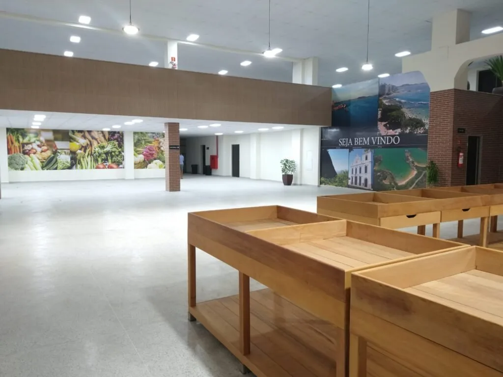 Interior de mercado com bancadas de madeira vazias e painel 'Seja bem vindo' com fotos de praias