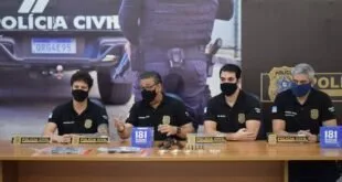 Policiais civis mascarados sentados atrás de mesa com armas e material apreendido