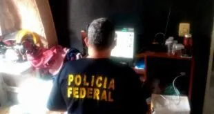 Agente de camiseta da Polícia Federal mexendo em um computador dentro de uma casa
