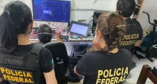 Agentes com coletes da Polícia Federal examinando computadores durante busca em residência