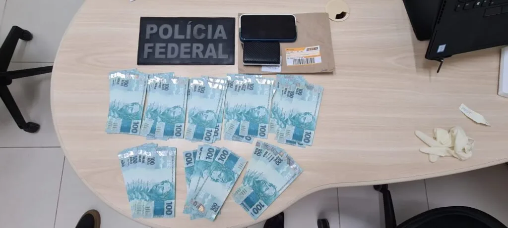 Cédulas de cem reais espalhadas sobre mesa ao lado de distintivo da Polícia Federal e um envelope