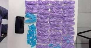 Dezenas de sacos e pinos de cocaína dispostos sobre superfície branca ao lado de um celular
