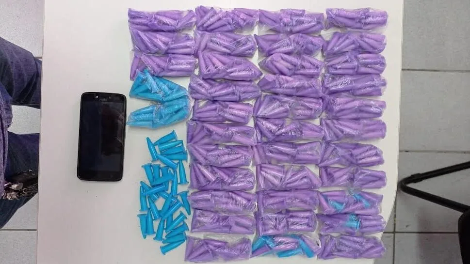 Dezenas de sacos e pinos de cocaína dispostos sobre superfície branca ao lado de um celular