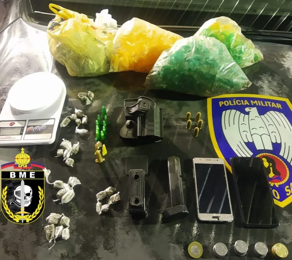 Sacos com entorpecentes, balança e celular apreendidos sobre mesa com brasão da Polícia Militar