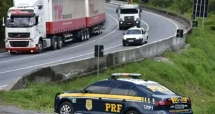 Viatura da PRF parada no acostamento de rodovia com caminhões trafegando ao fundo