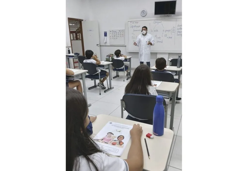 Alunos de máscara sentados em carteiras enquanto professor de jaleco escreve no quadro branco