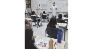 Alunos de máscara sentados em carteiras enquanto professor de jaleco escreve no quadro branco