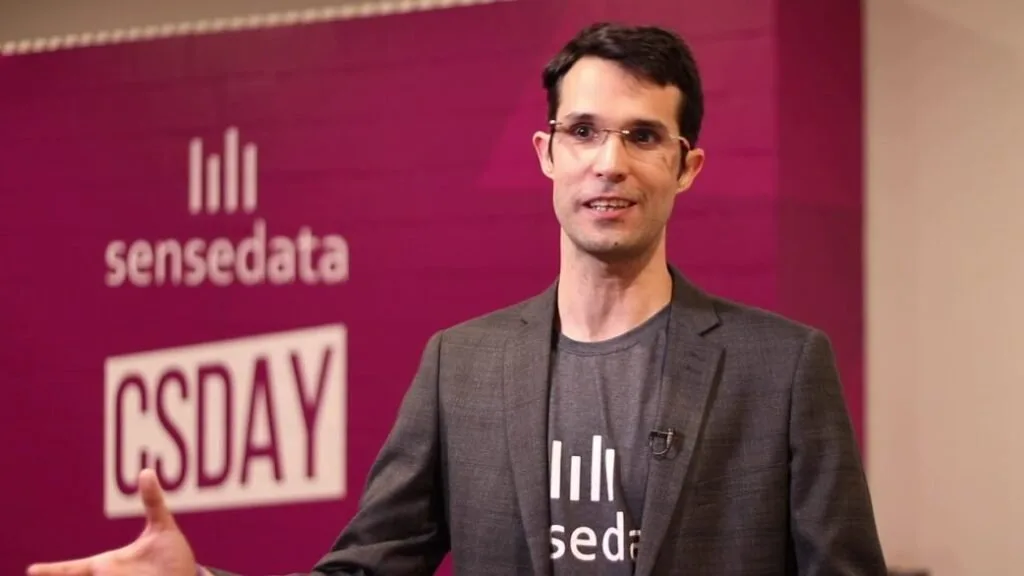 Homem de óculos e camiseta preta fala em evento diante de painel rosa com o logotipo SenseData